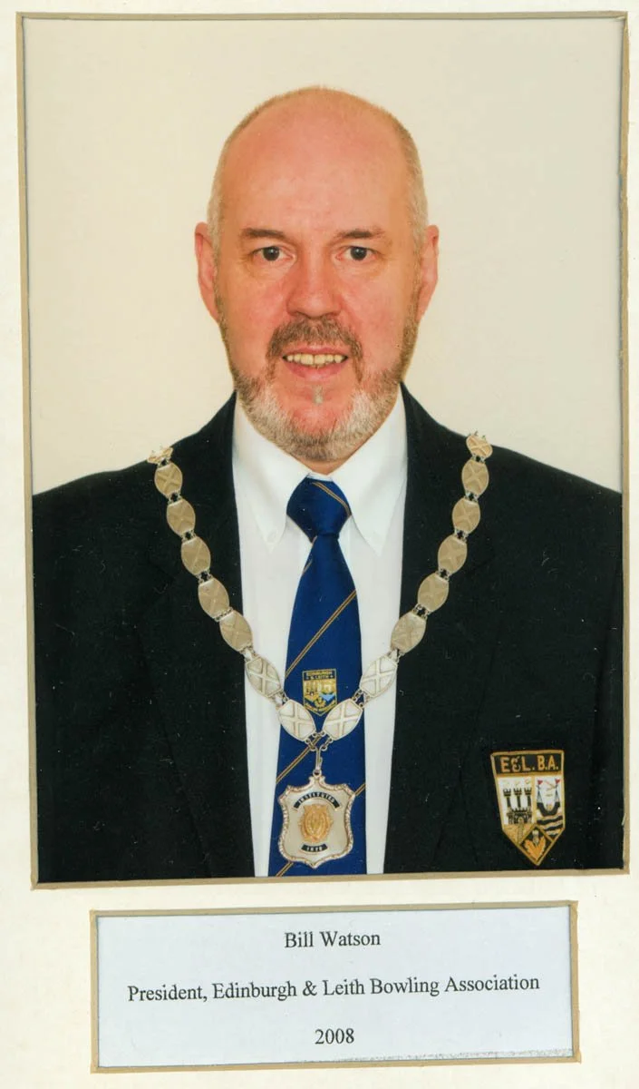 Photograph of Bill Watson wearing a suit, tie, and a chain of office with a badge, indicating he is a mayor or official, with a badge on his jacket and a plaque below bearing his name and title as President of Edinburgh & Leith Bowling Association in