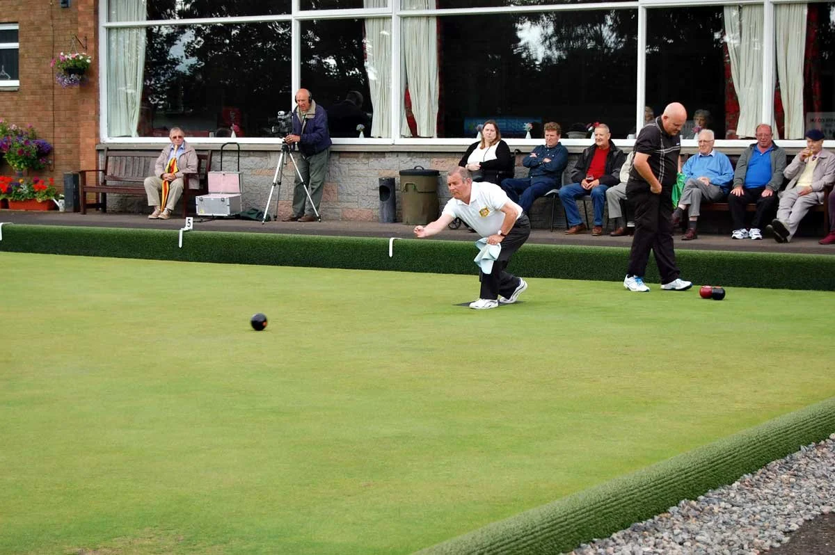 A group of people watching a lawn bowling game outdoors with a building and large windows behind them. Some spectators are seated on benches, while others are standing. One player is preparing to release the jack, and another player is standing nearb