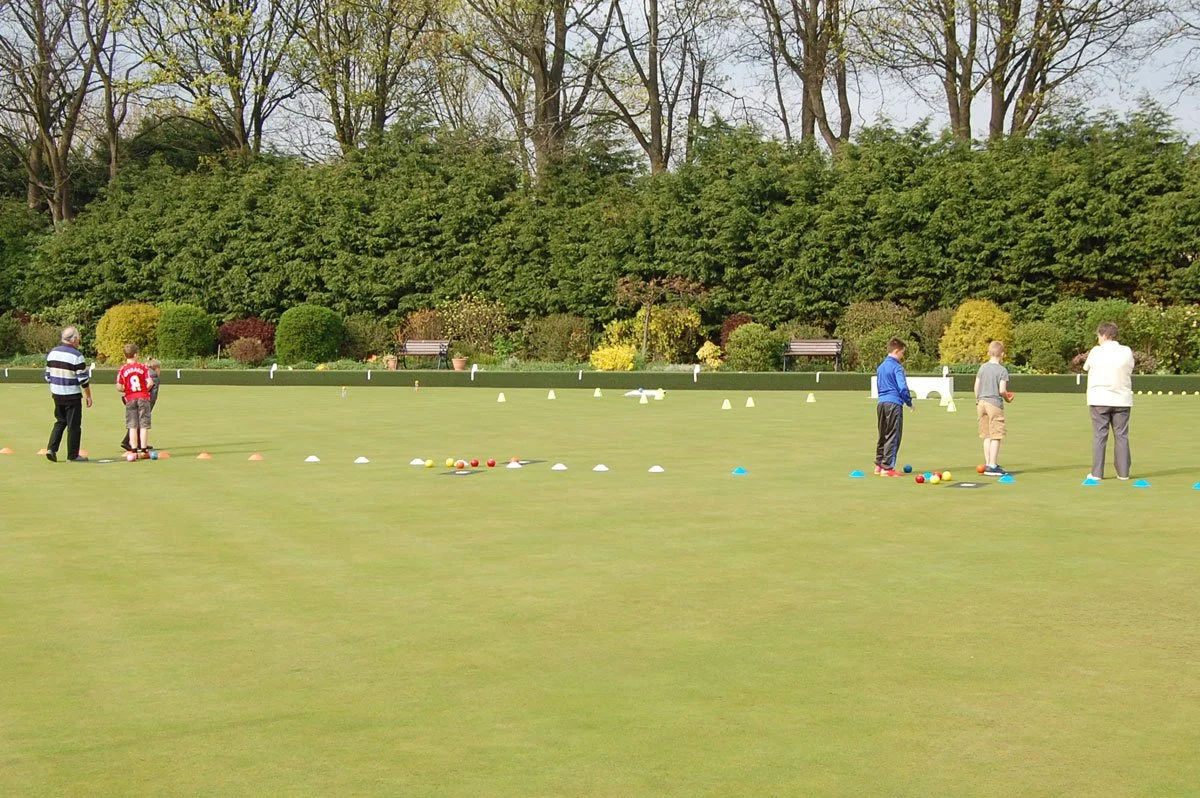 People playing lawn bowls or bocce on a green field with colorful balls and cones, surrounded by trees and benches.