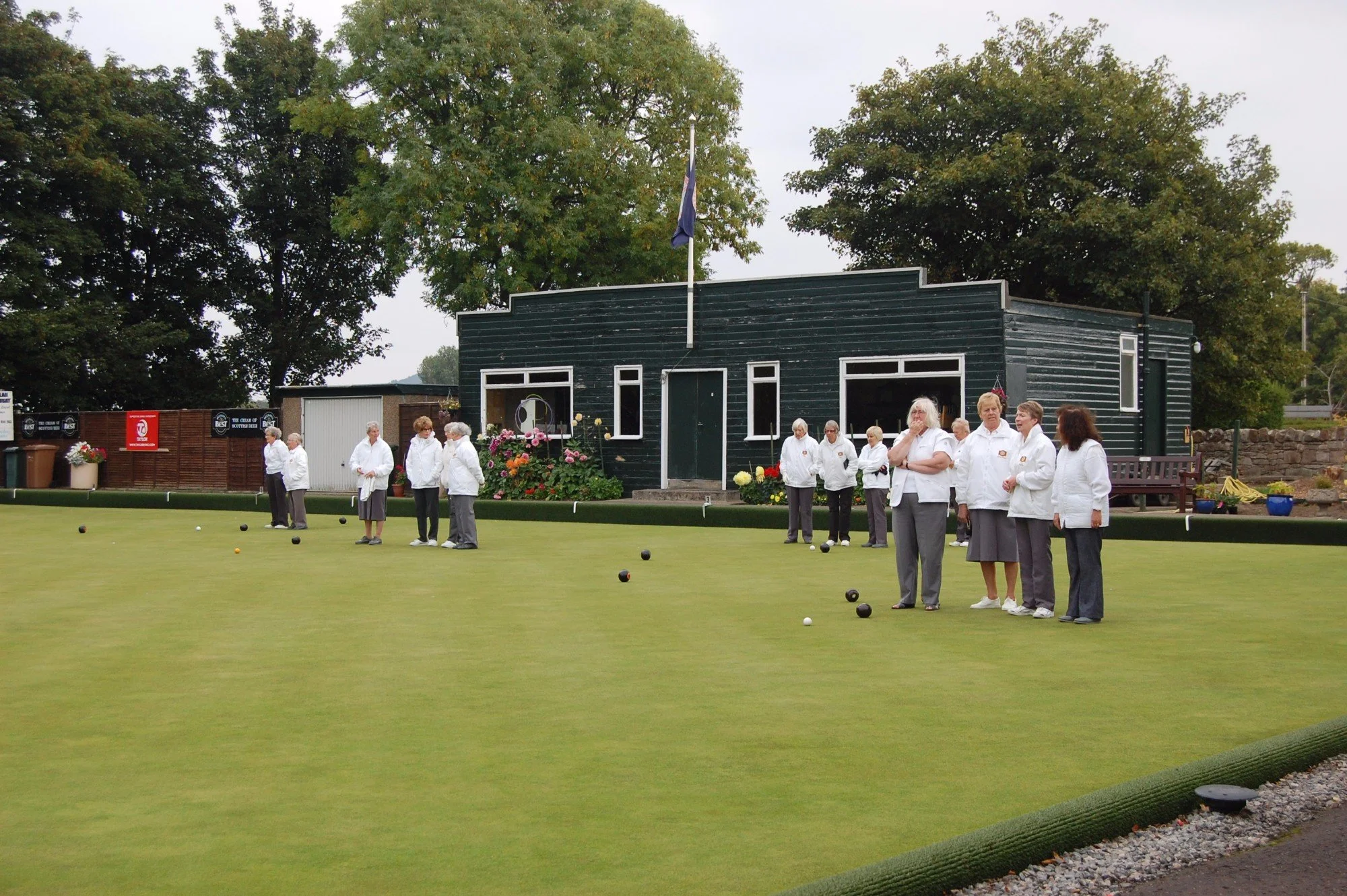 A group of mostly elderly women playing lawn bowls on a green field with a dark green wooden building in the background, surrounded by large trees.