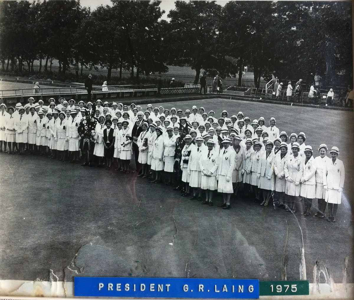 Black and white photograph of a large group of women dressed in white coats and hats, standing in formation on a field, with a few men in uniform among them. In the background, there are trees, a fence, and more women in white coats. A blue label at 