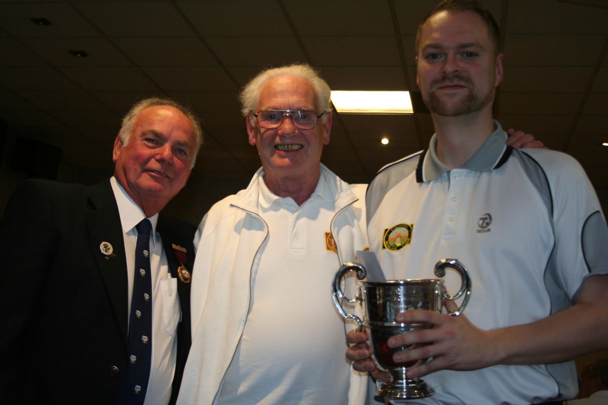 Three men in formal and sports clothing posing indoors, with the man on the right holding a trophy.