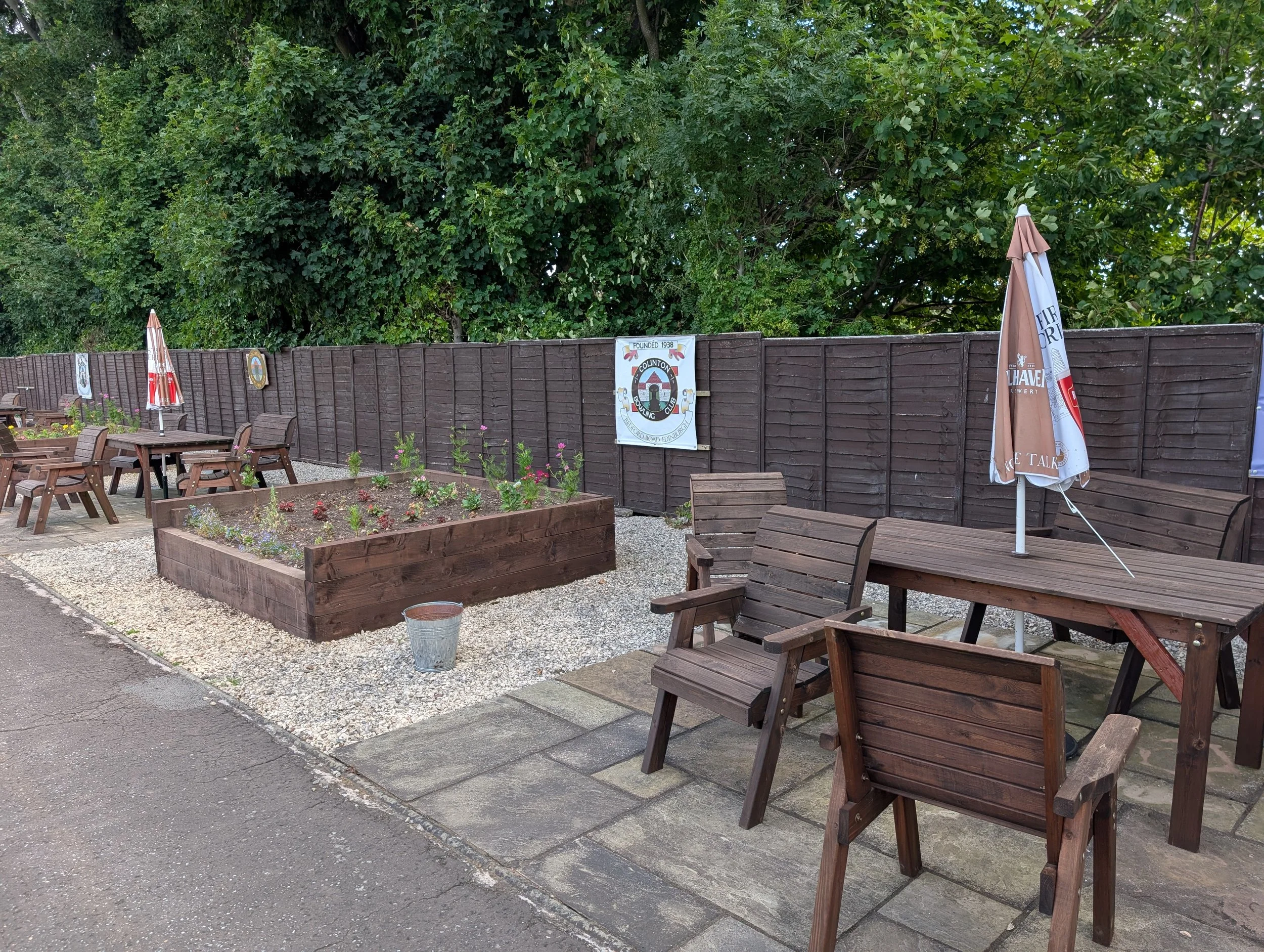Outdoor patio area with wooden chairs, flower beds, a picnic table with an umbrella, and a wooden fence with a logo sign, set against a backdrop of lush green trees.