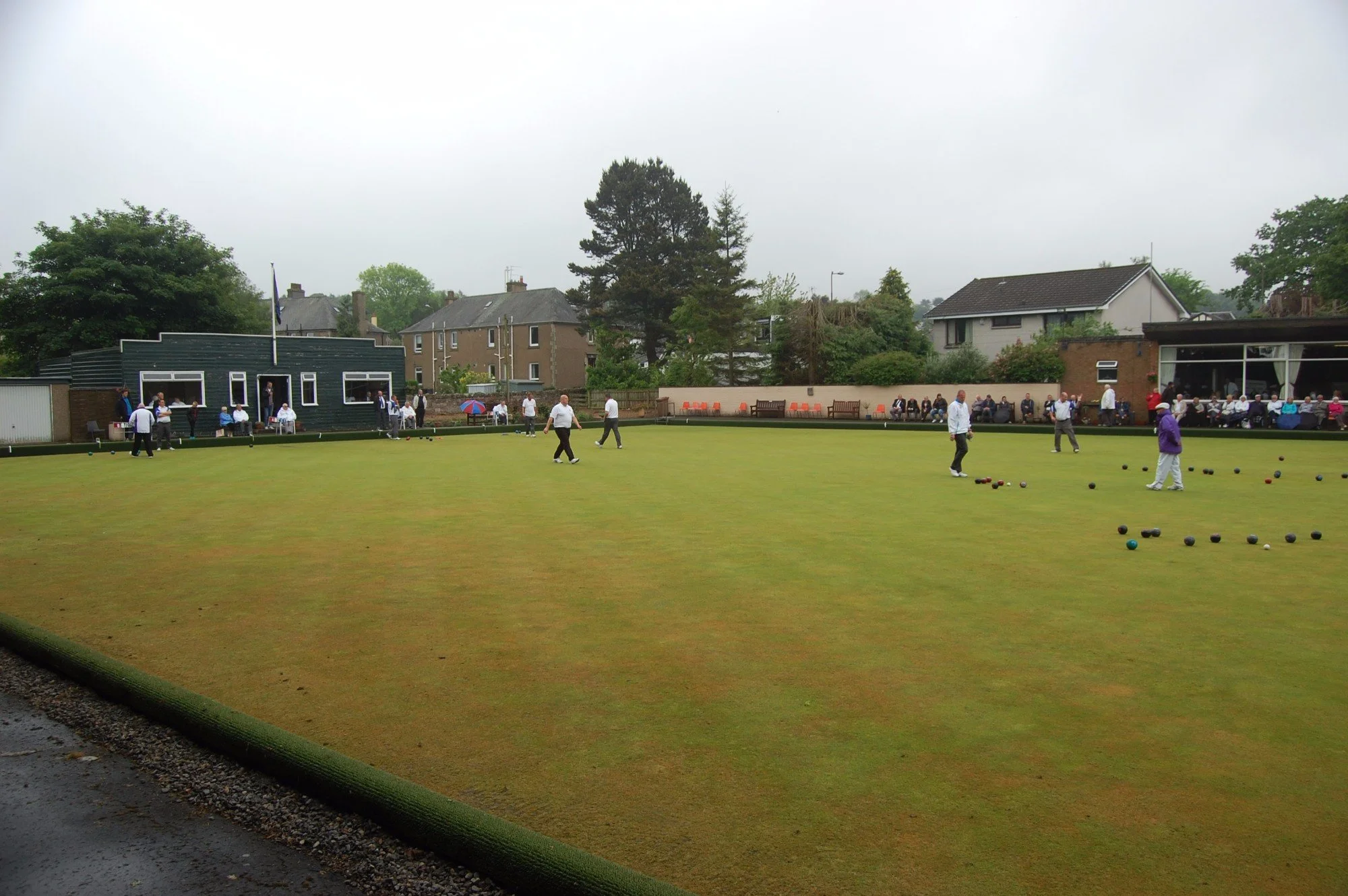 People playing lawn bowls on a green field with spectators seated nearby and residential buildings in the background.