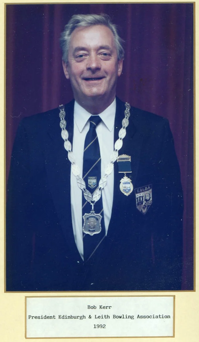 A man in a suit wearing medals and badges, standing in front of a red curtain, with a label below that reads: Bob Kerr, President Edinburgh & Leith Bowling Association, 1992.