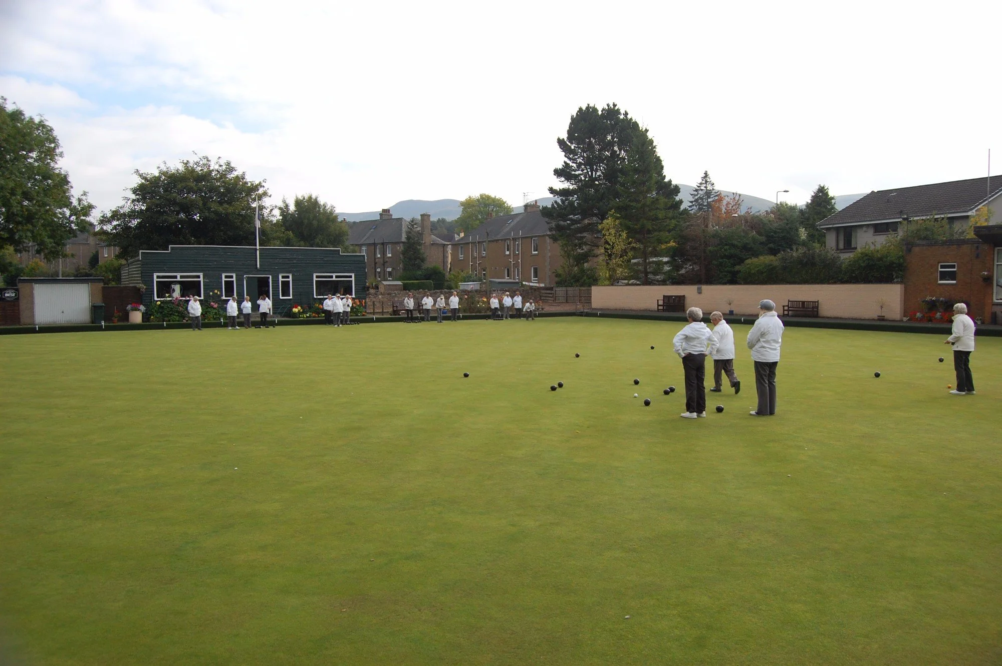 A group of people dressed in white playing the game of bowls on a well-maintained green lawn, with houses, trees, and hills in the background.
