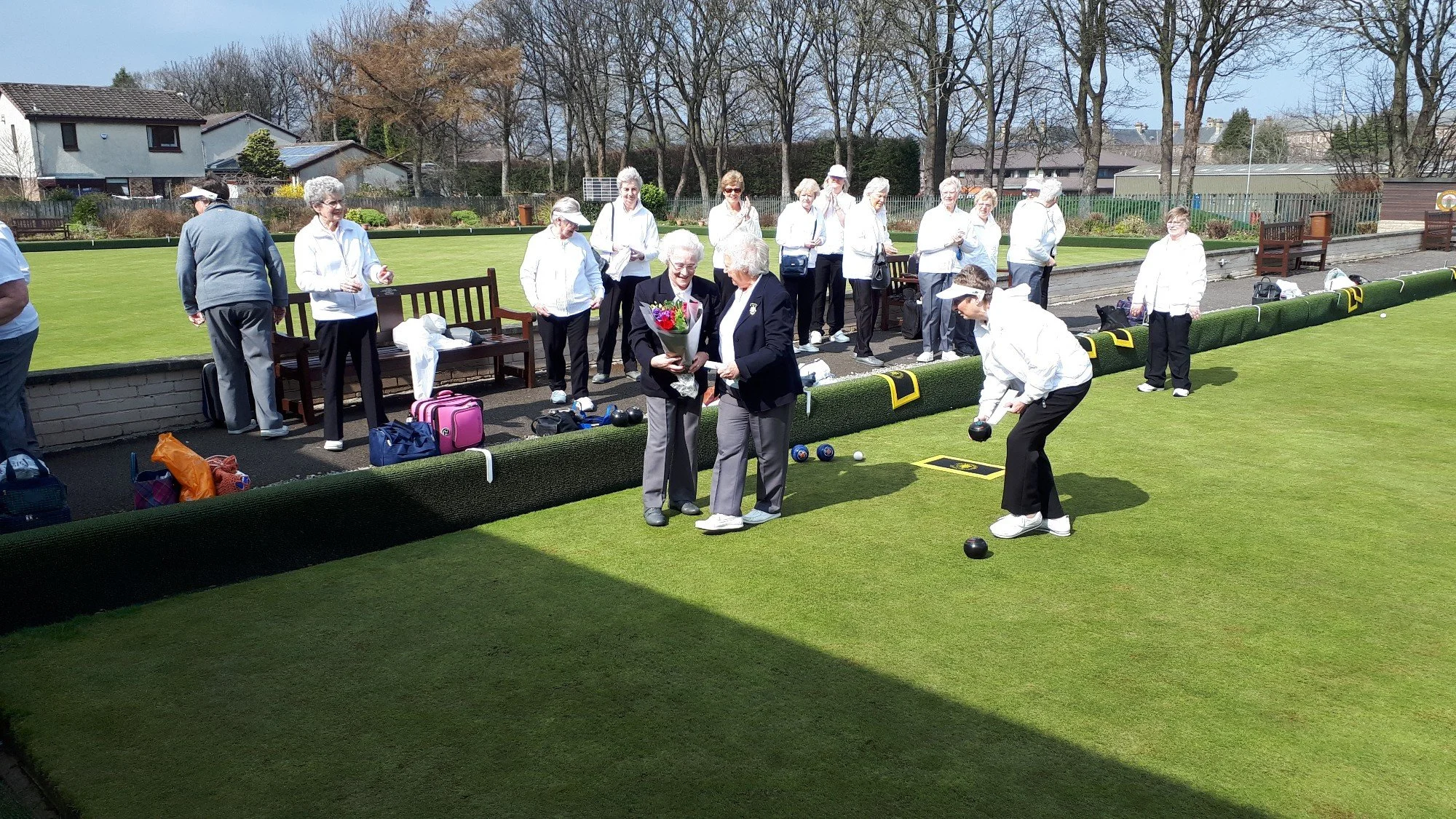 Group of elderly women outdoors on a lawn playing lawn bowls, some watching, others preparing to throw bowls, with benches, trees, and houses in the background on a sunny day.