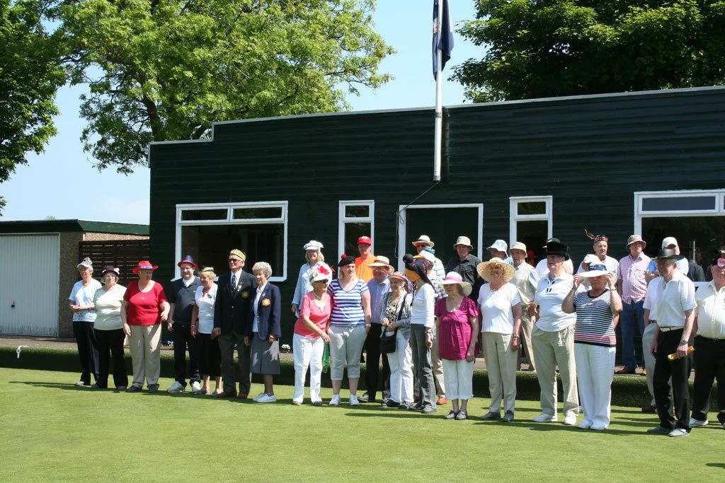 Group of mostly senior adults standing on a lawn in front of a black building, dressed in summer clothing and hats, some wearing sunglasses, under clear weather with trees in the background.
