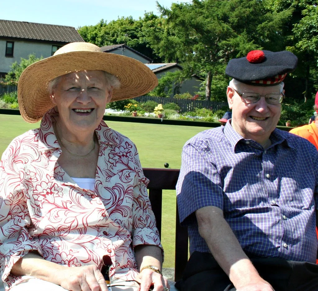 An elderly woman and man sitting outdoors, smiling. The woman is wearing a large straw hat and a patterned blouse, holding a walking stick. The man has glasses and is wearing a black hat with a red pom-pom and a purple checkered shirt. Green trees an