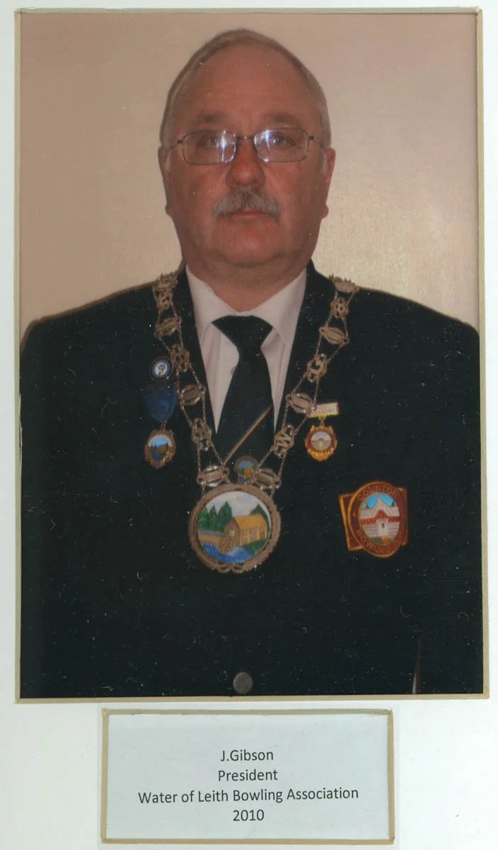 A man wearing glasses, a dark suit, tie, and a chain with large medallions, standing against a plain background. There is a nameplate that identifies him as J. Gibson, President of the Water of Leith Bowling Association in 2010.
