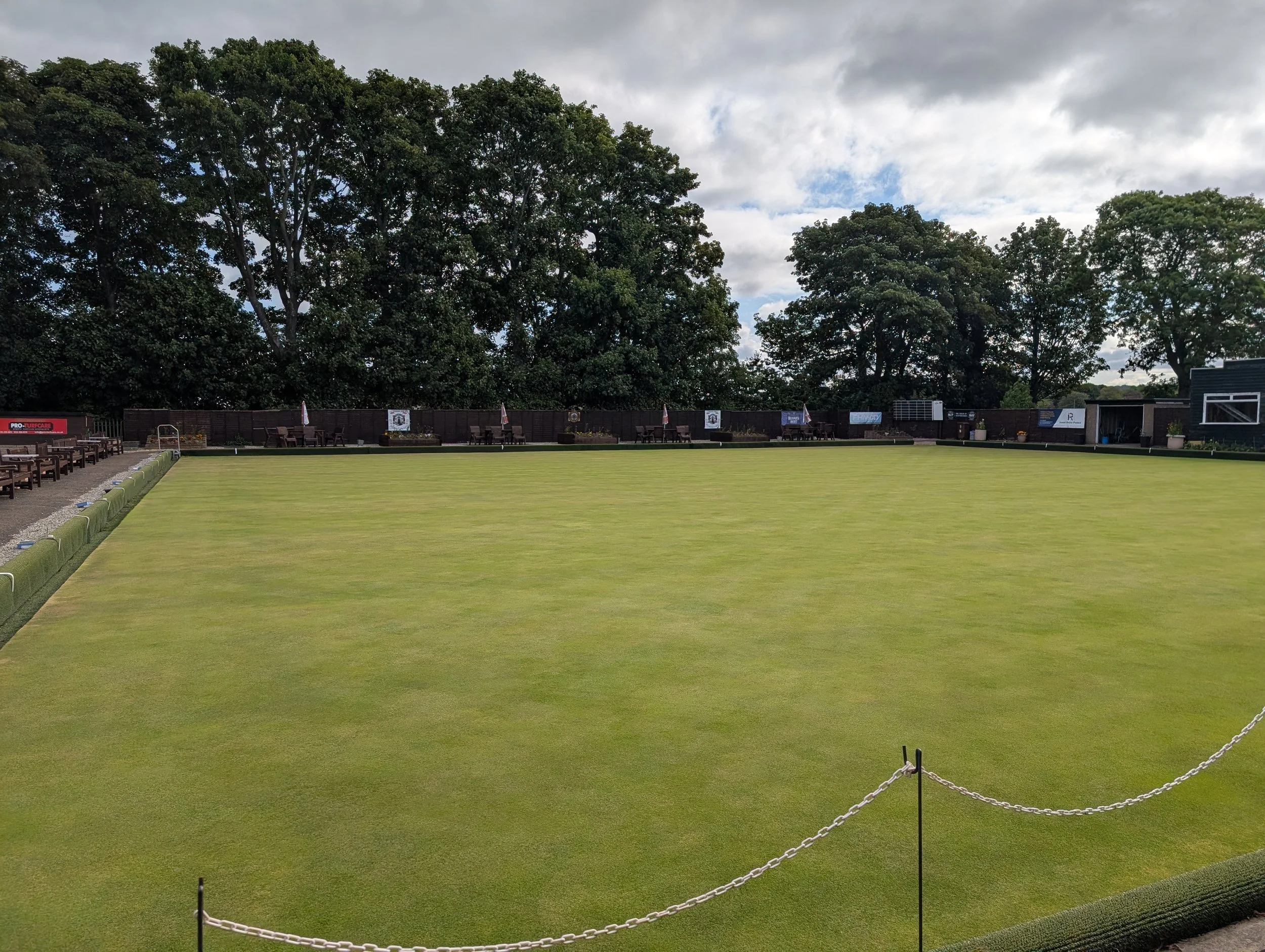 A green outdoor lawn bowling green with benches around it, surrounded by a low chain fence, and tall trees in the background under a partly cloudy sky.