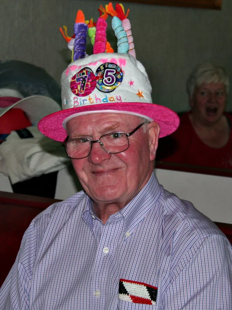 Senior man wearing a colorful birthday hat with candles and numbers, smiling and dressed in a striped shirt, in a festive setting with a woman in the background.