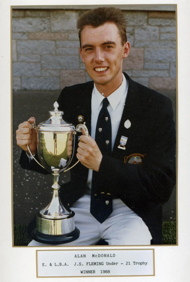 A young man in a black blazer, white shirt, and dark tie holding a large silver trophy, sitting outdoors against a brick wall. He is smiling and wearing pins on his blazer, with a patch on his sleeve. The caption below states his name as Alan McDonal