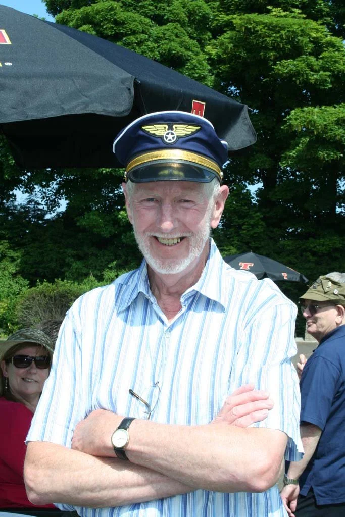 An elderly man wearing a pilot's hat with wings and a star emblem standing outdoors with a smile, crossed arms, and a striped shirt, with people and umbrellas in the background.