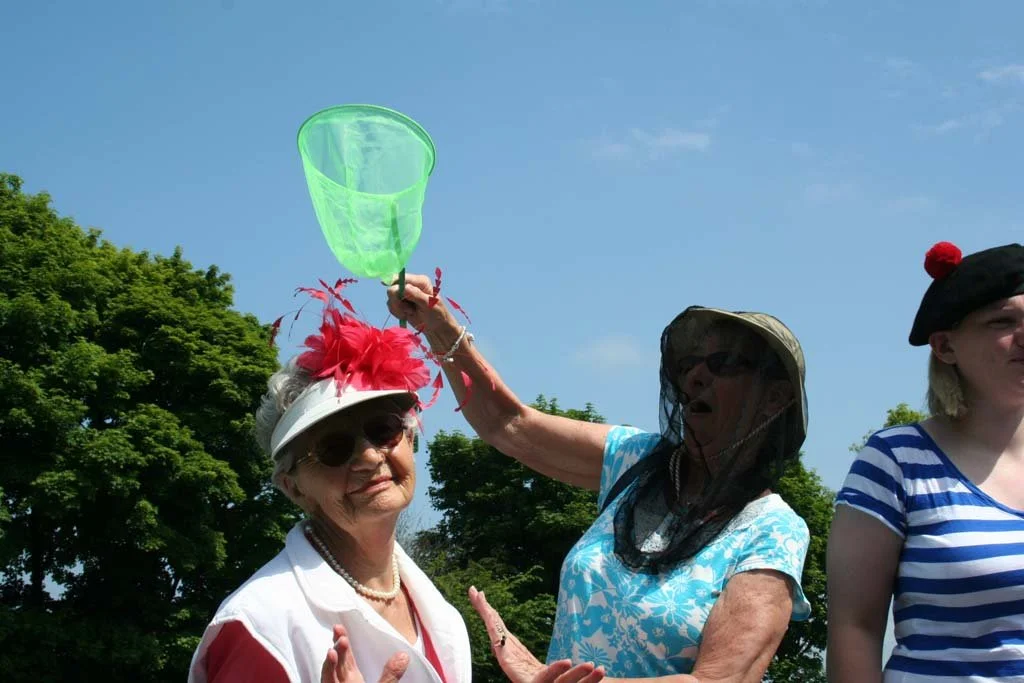 Three women outdoors, two wearing sun hats and sunglasses, one woman holding a green butterfly net above the head of an older woman.