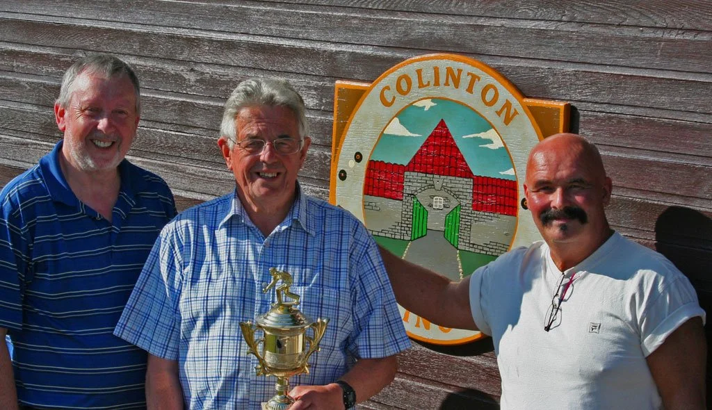 Three men standing in front of a building with a wooden exterior, holding a trophy. The man in the middle wears a checkered shirt and glasses, while the man on the right has a mustache and is in a white shirt. The man on the left is in a blue striped