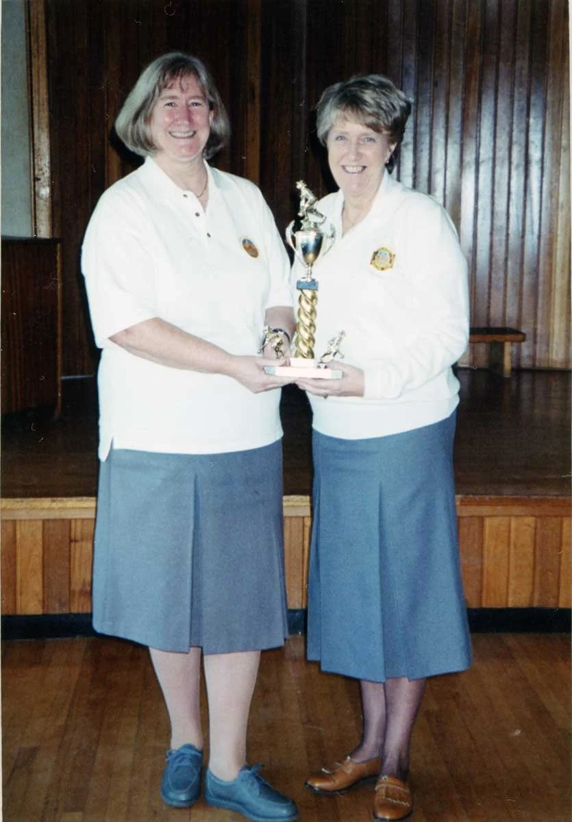 Two women in matching white jackets and gray skirts standing on a wooden floor, smiling, holding a trophy together in a room with wood-paneled walls.