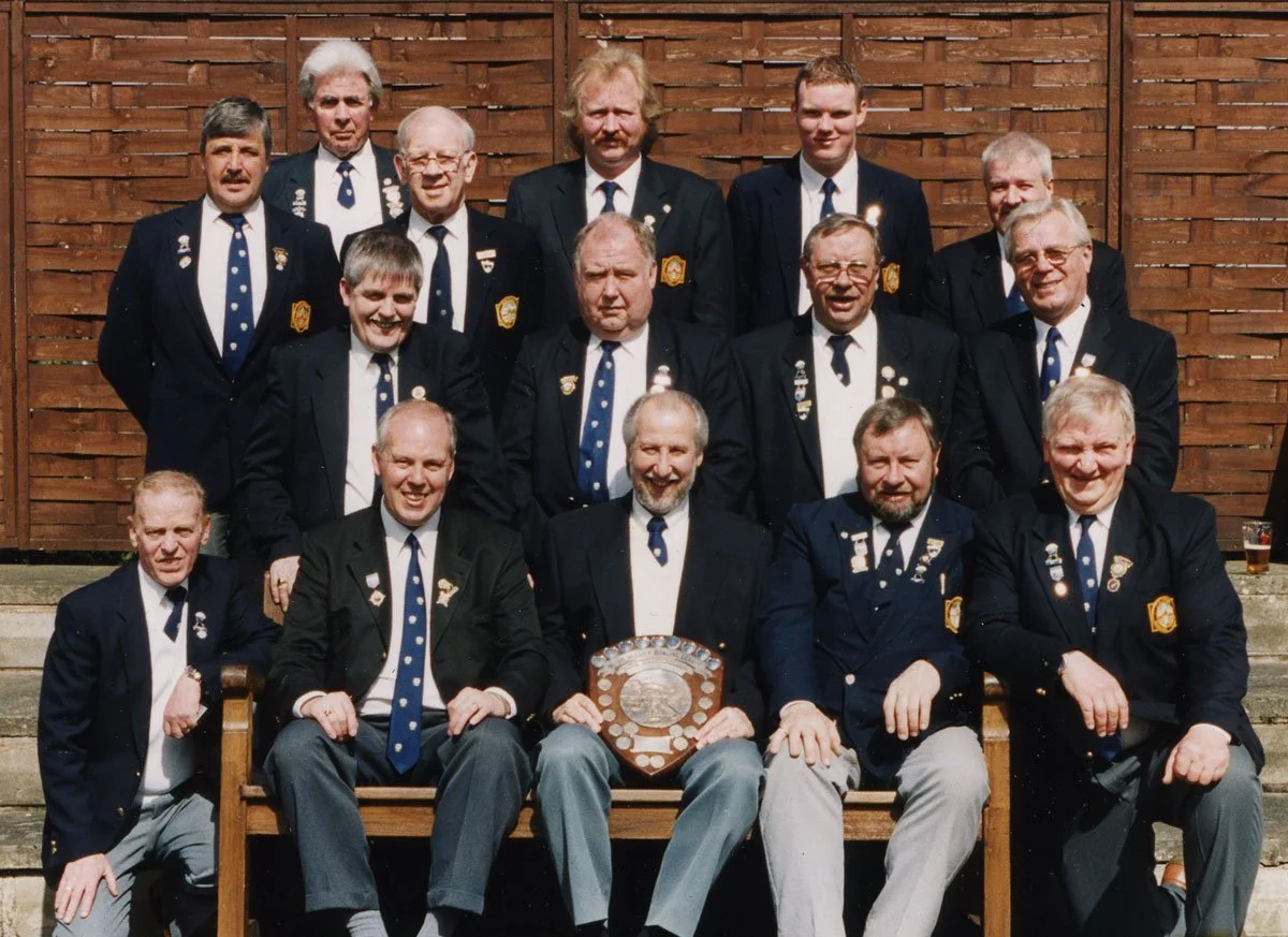 Group of men dressed in blue blazers with pins and badges, posing on steps in front of a wooden fence. One man holds a large shield-shaped plaque. They are smiling, likely at a formal gathering or award event.