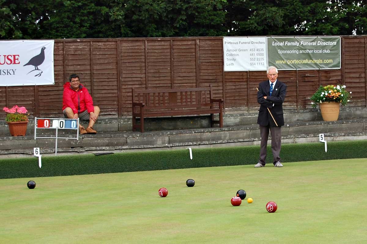 Two men playing lawn bowls on a green, with a wooden bench and flower pots in the background. One man stands with arms crossed, wearing a suit, and the other sits on a bench wearing a red jacket. Two signs with scores are visible.