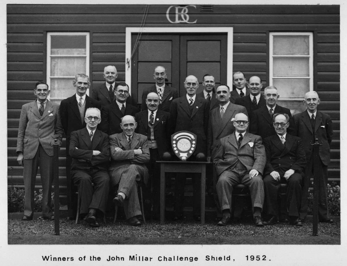 Black and white photo of 15 men in suits outdoors, with a building in the background. The men are posing for a group photo, with some seated and others standing. There is a shield with the John Millar Challenge logo on a table in front.