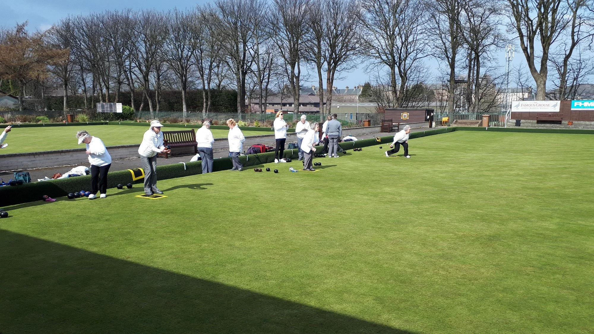 People playing lawn bowls on a green field on a sunny day with trees in the background.