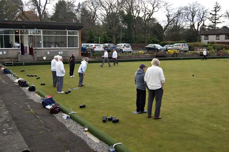 People playing lawn bowls on a grass field, with bags and bowls on the ground, and a building with glass windows in the background.