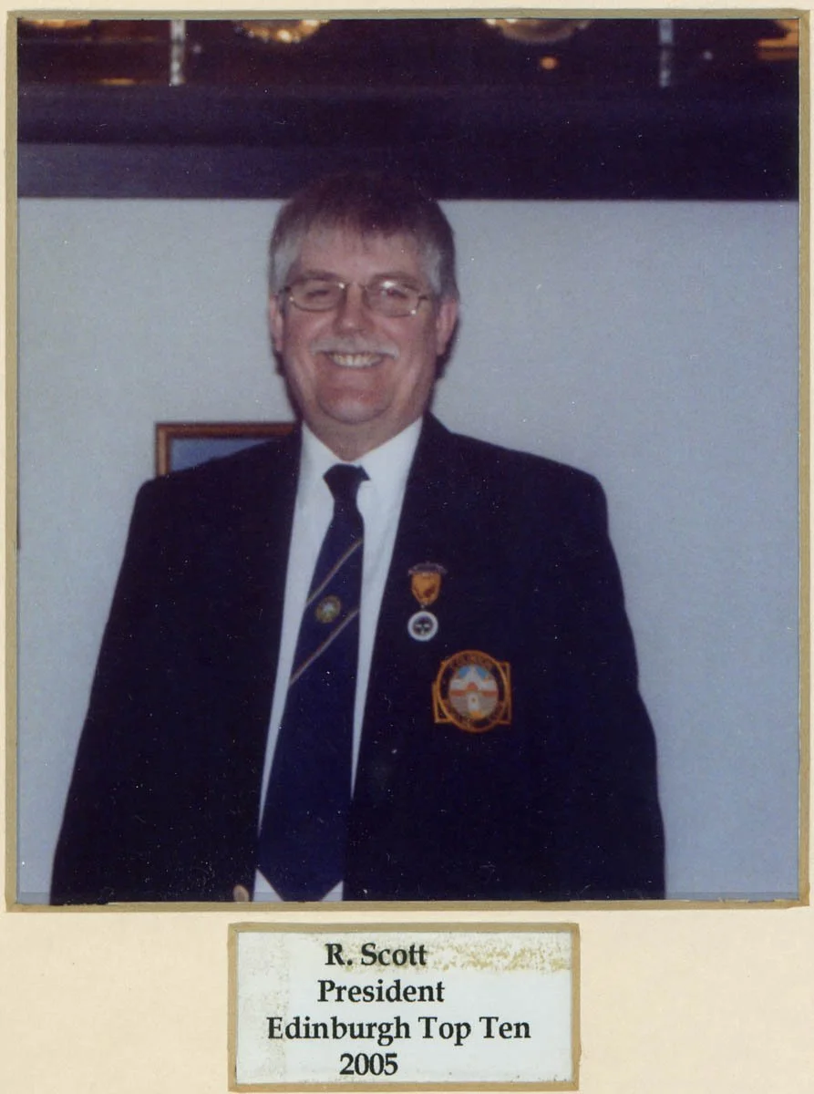Photograph of R. Scott wearing a suit and tie with pins, smiling, in a room with a framed picture on the wall, identified as President of Edinburgh Top Ten in 2005.