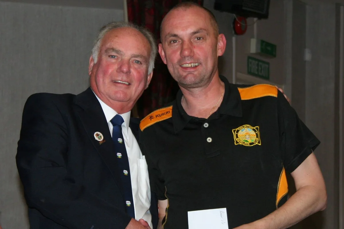 Two men standing side by side and smiling, one in a black suit and the other in a black parking enforcement shirt with orange accents, shaking hands at an indoor event.
