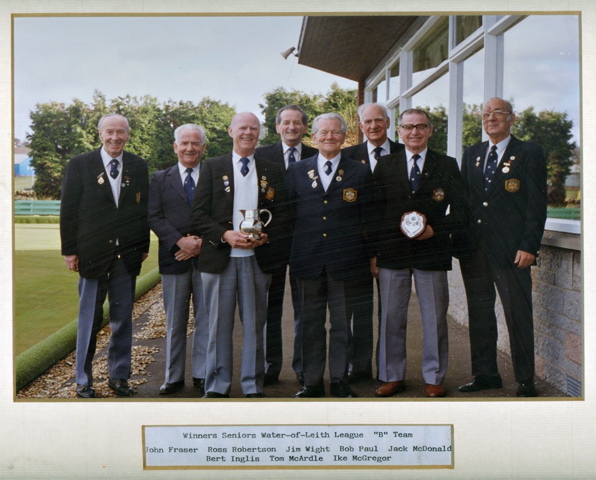 Group of eight senior men dressed in blazers with medals, standing outdoors with greenery and a building in the background, holding trophies. Caption indicates they are winners of the Seniors Water-of-Leith League 'B' Team.