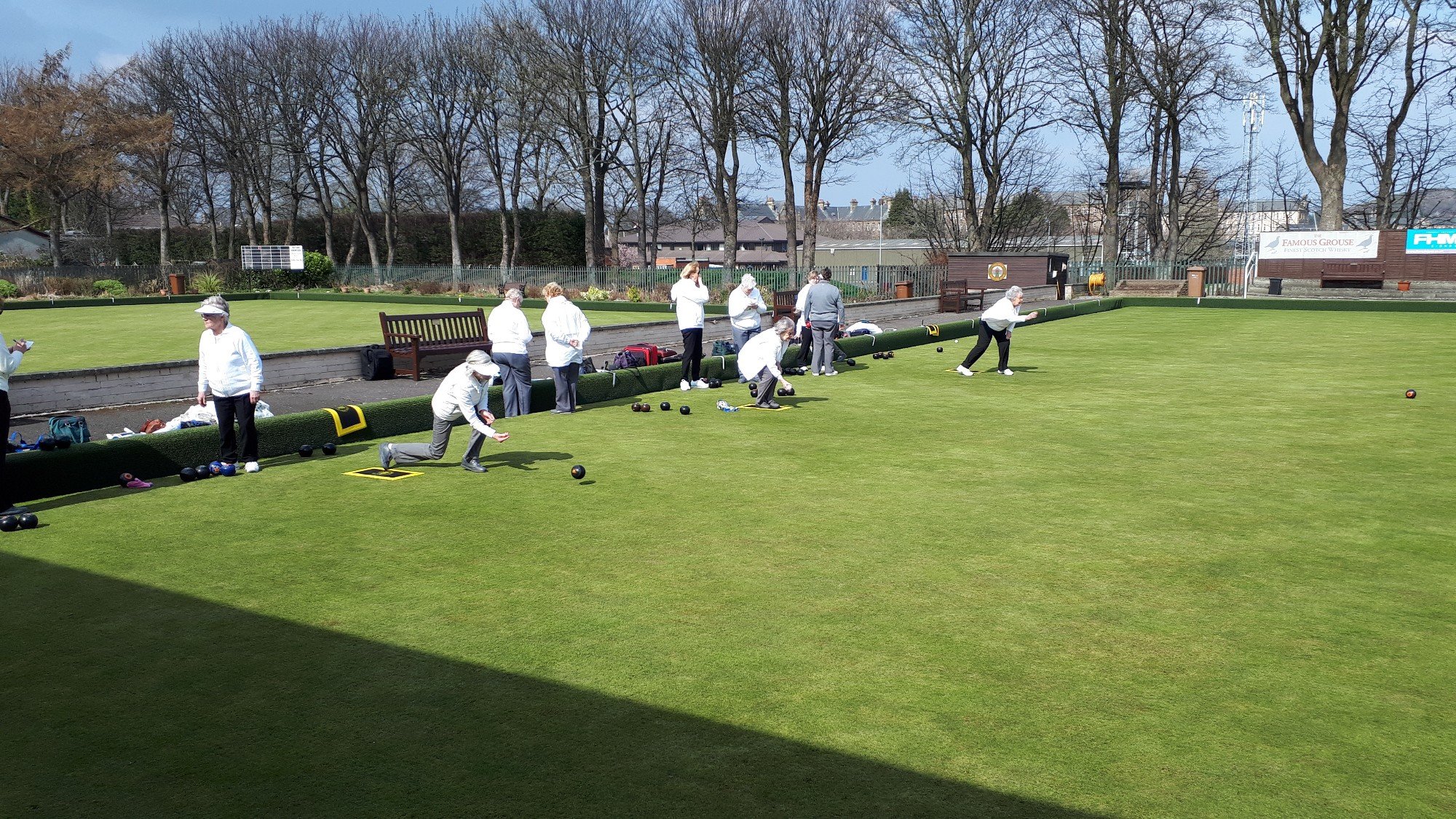 A group of people playing lawn bowls on a well-maintained grassy field, some are participating while others observe, with trees and signs in the background on a sunny day.