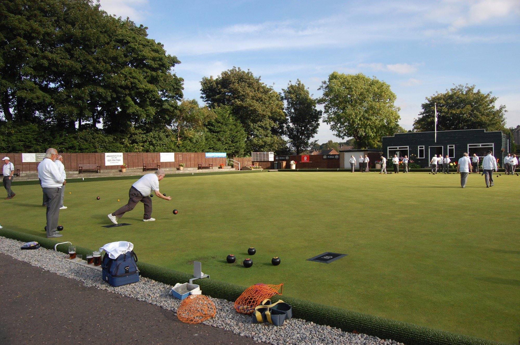 People playing lawn bowls on a manicured green field with trees and a dark building in the background, and various bowls, bags, and drinks on the side.