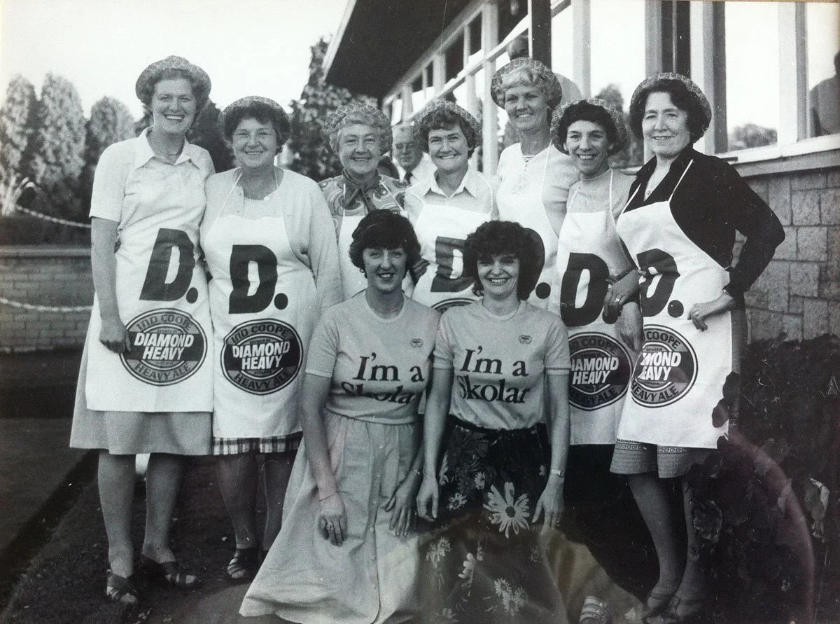 Ten women posing outdoors, some wearing aprons with slogans, in front of a building with windows. The women are smiling and standing in a group.