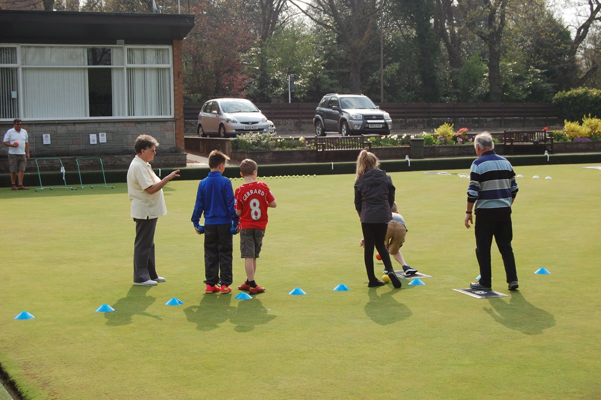 Group of children and adults playing lawn bowls on a grass field, with cones marking the playing area and equipment in the background.
