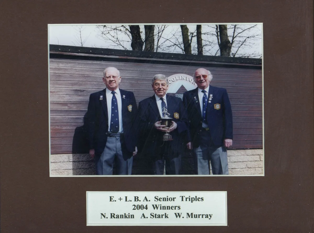 Three elderly men in suits with pins standing outdoors in front of a wooden wall, holding a trophy, with a sign that reads "COUNTY" behind them, commemorating the 2004 winners of E. + L. B. A. Senior Triples, N. Rankin, A. Stark, and W. Murray.