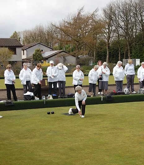 A group of elderly women playing lawn bowls on a green, with some preparing to bowl and others watching, in an outdoor park setting.