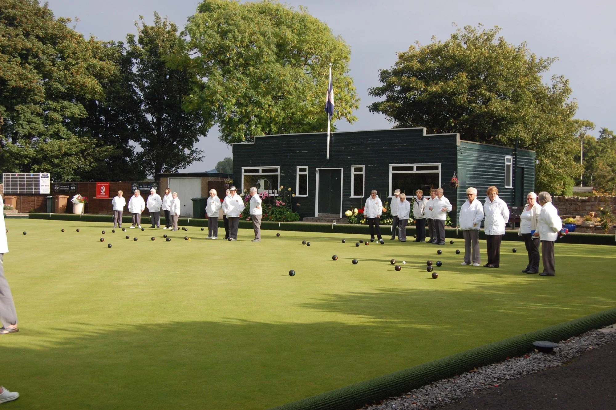 A group of elderly women in white jackets playing lawn bowls on a green field, with large trees and a building with a flag in the background.