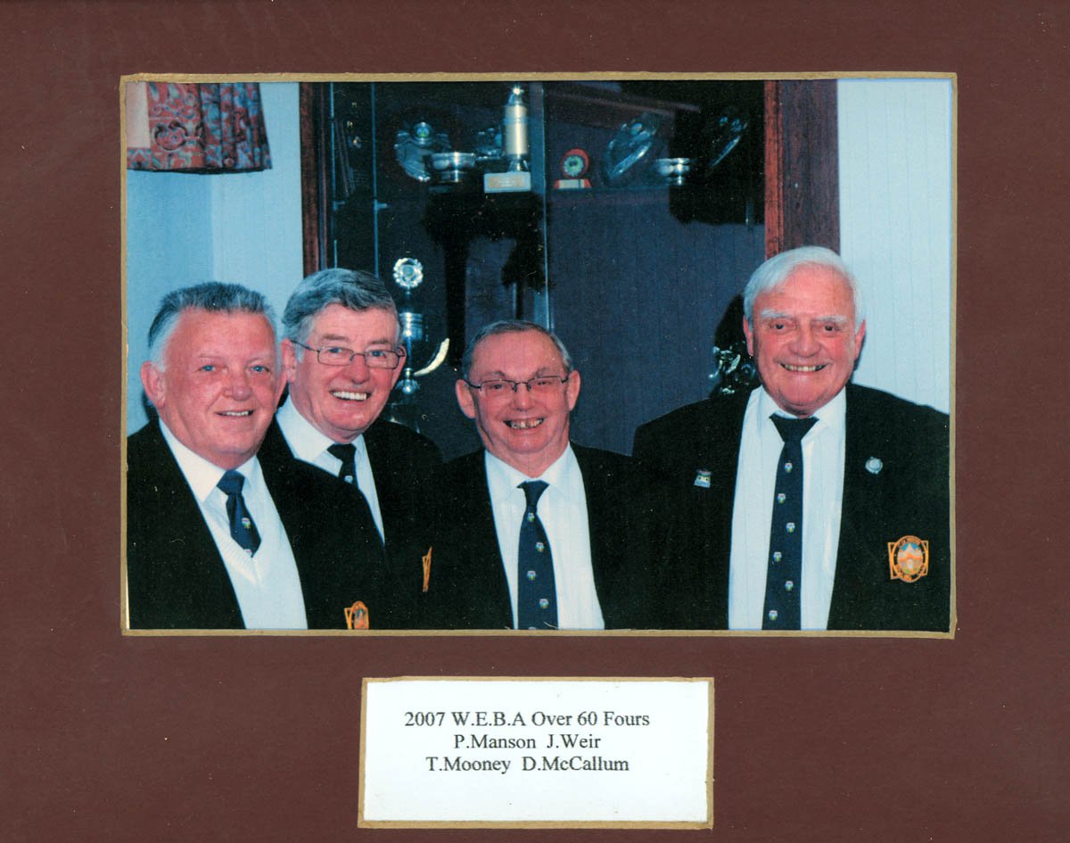 Four older men dressed in navy blue blazers and white shirts, smiling for a photo, in a display frame with a note below. The note reads: "2007 W.E.B.A Over 60 Fours P. Manson J. Weir T. Mooney D. McCallum."