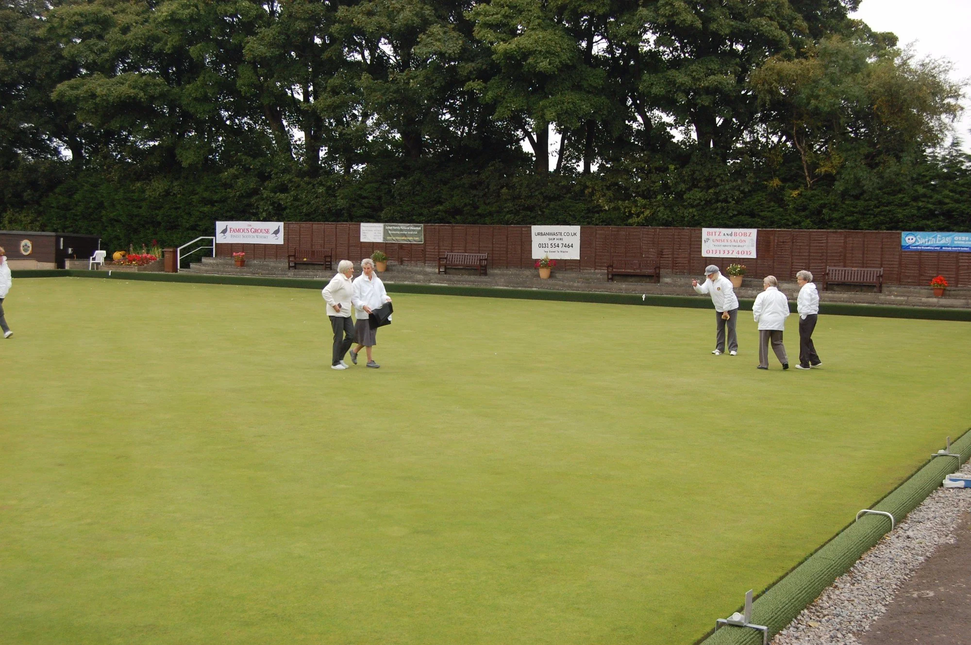 A lawn bowls green with five elderly women, four of whom are dressed in white, standing and chatting. Six additional women are further away on the green, with more women also nearby. There are advertisement banners along a wooden fence in the backgro
