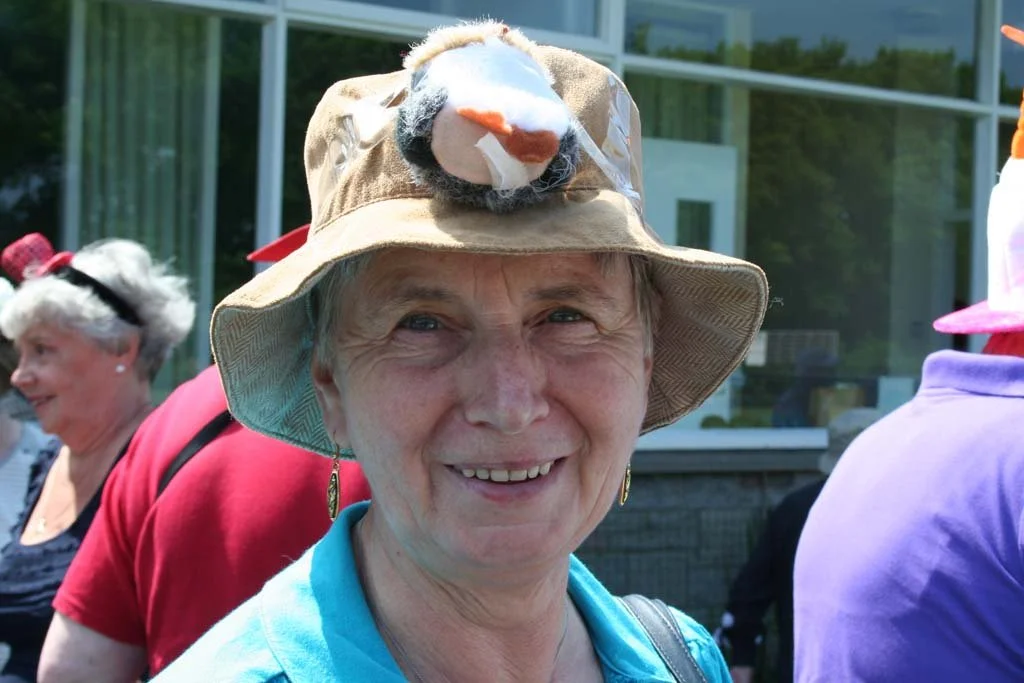 A smiling elderly woman wearing a tan hat with a plush tiger face on top, outdoors with other people in the background.