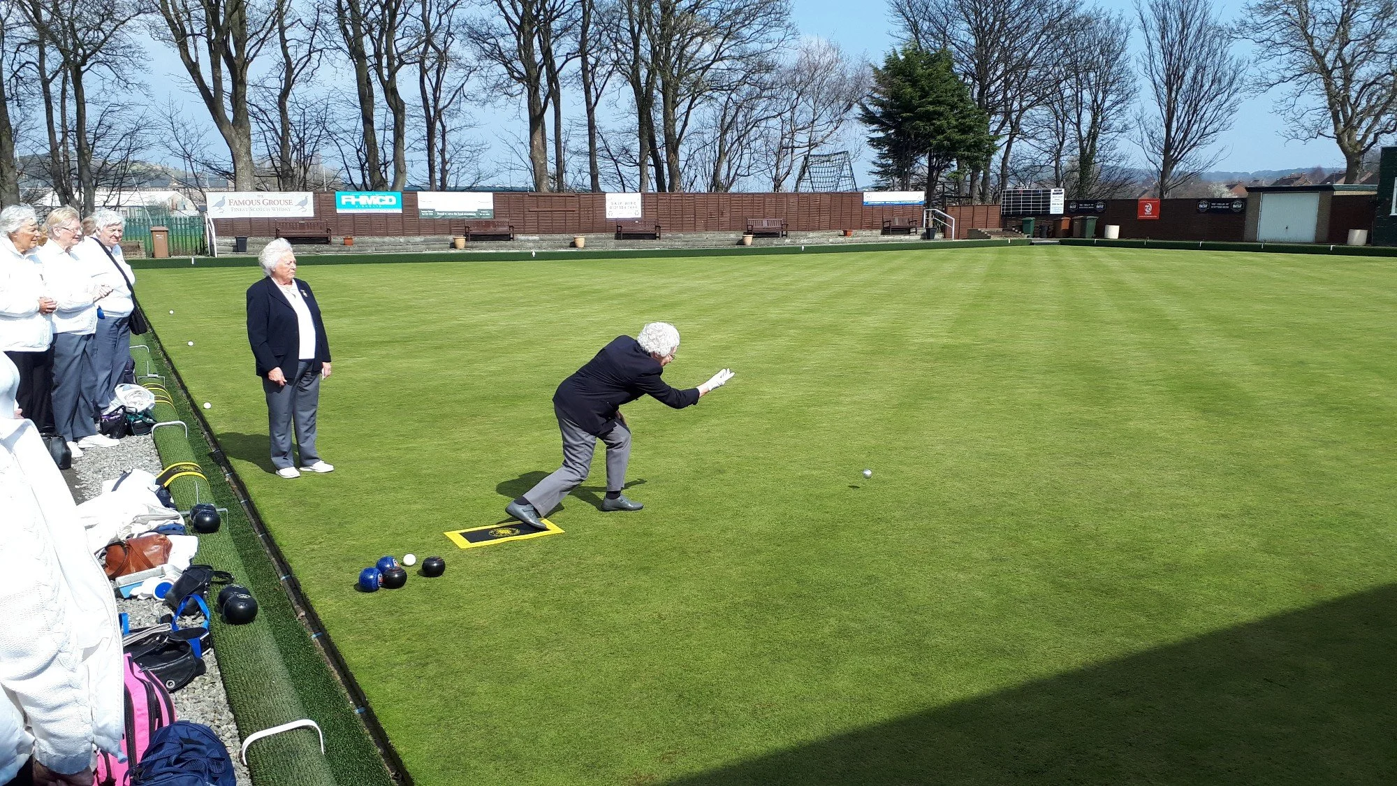 A group of elderly women watching a man in a blazer and gloves rolling a curling stone on a grass field, with curling stones and bags on the ground nearby, and trees with bare branches in the background.