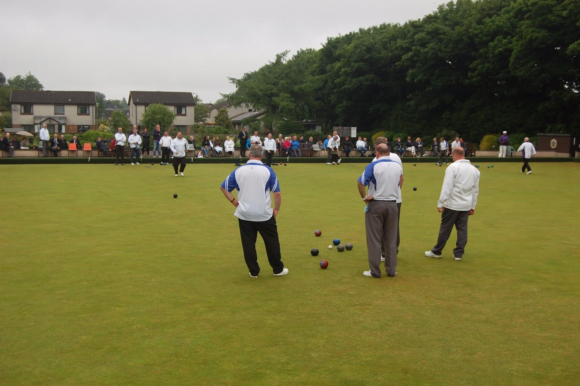 People playing lawn bowls on a green, with spectators sitting on benches in the background.