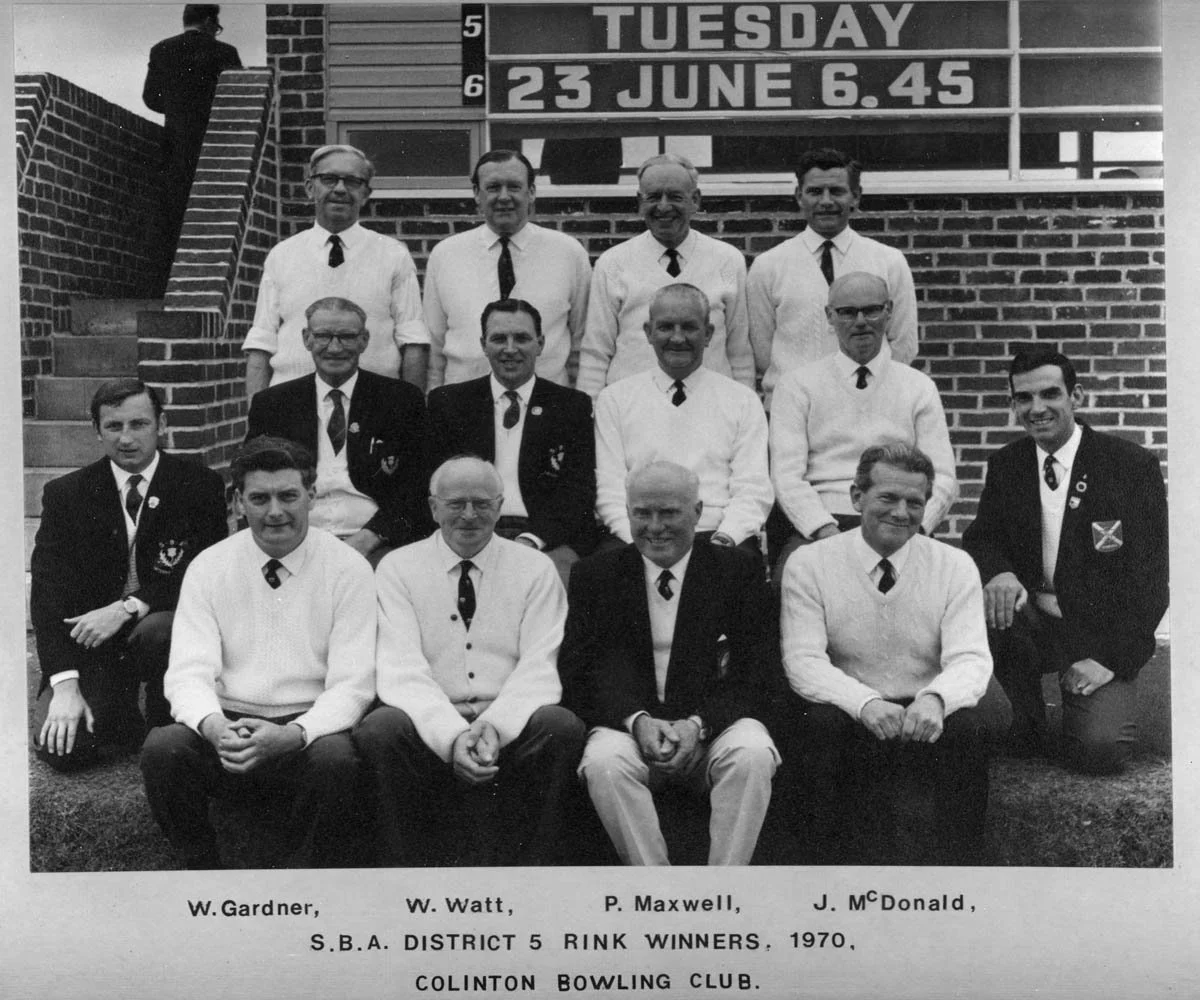 Black and white photo of fourteen men posing in front of a brick building and an outdoor sign. The sign shows 'Tuesday, 23 June 6.45'. A few men are wearing suits with ties, some wearing white sweaters, and some in blazers with a logo. The bottom tex