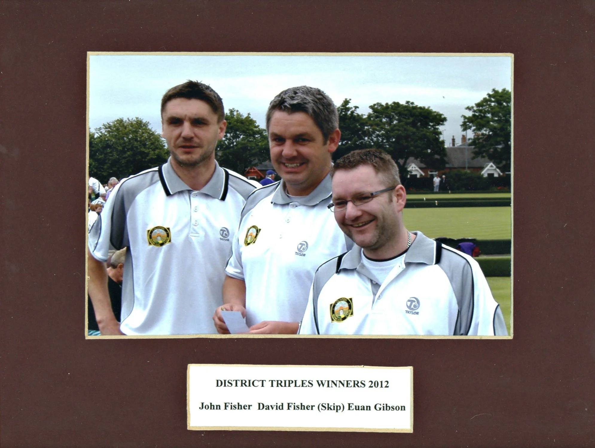 Three men in matching sports uniforms standing outdoors on a sports field with trees and houses in the background, smiling for a photo. The caption indicates they are district triples winners 2012, named John Fisher, David Fisher (Skip), and Euan Gib