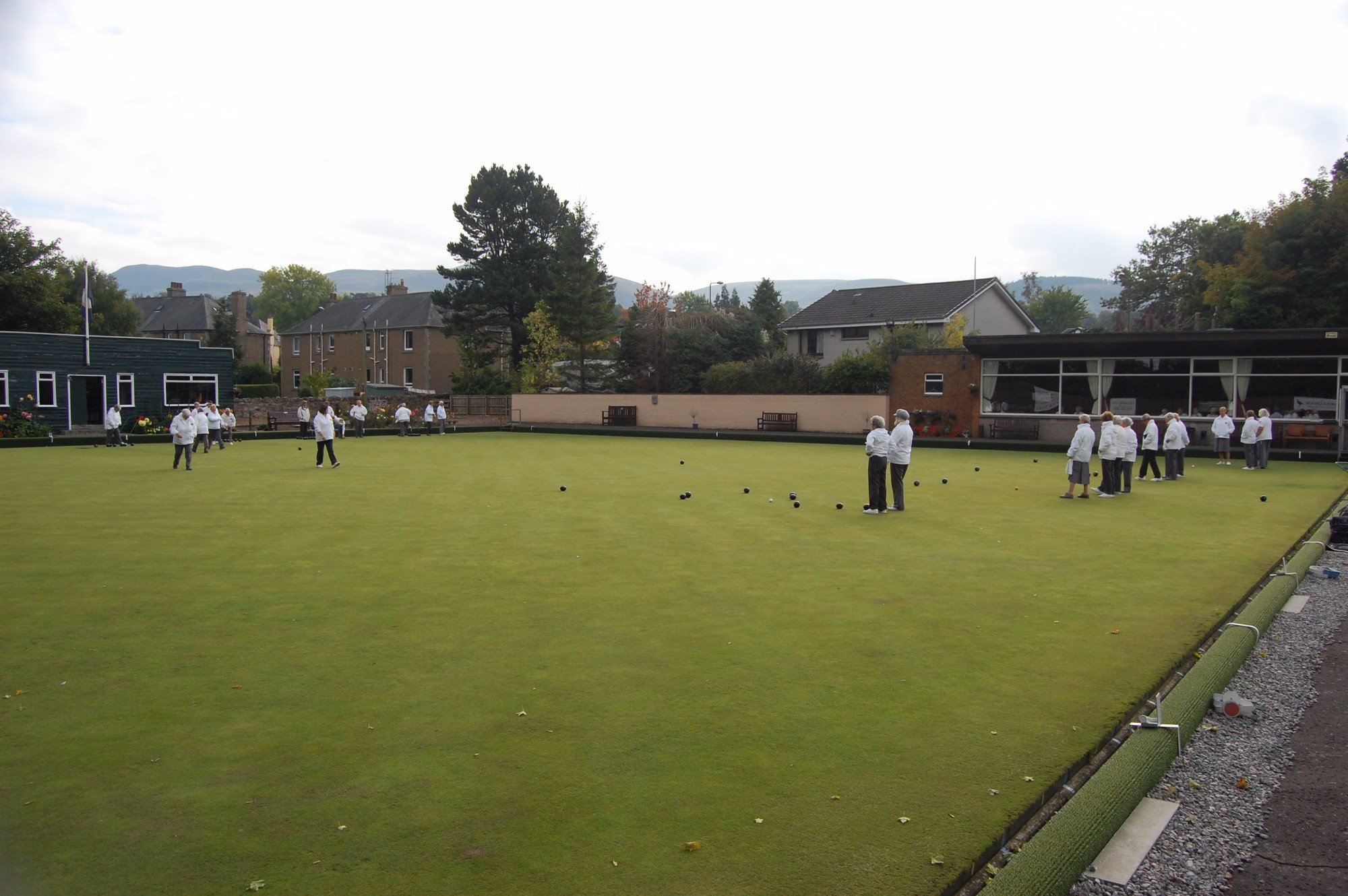 People playing lawn bowls on a green surrounded by trees and residential houses, with mountains in the background.