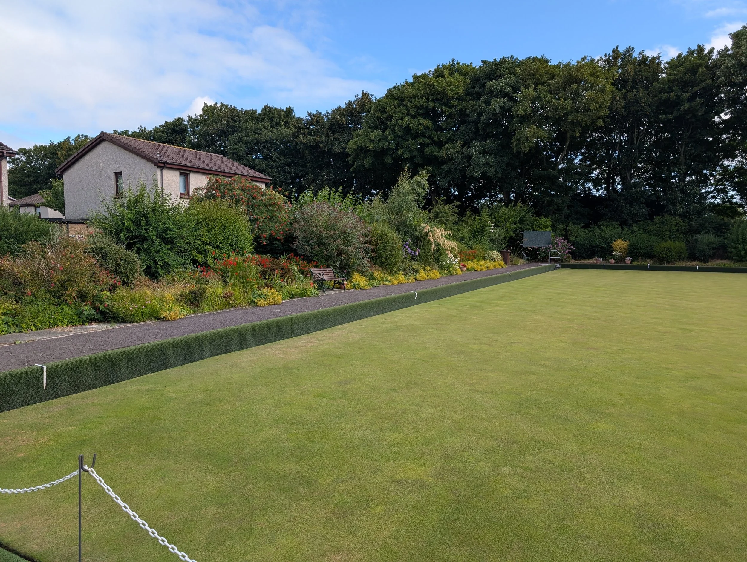 A well-maintained lawn with a chained fence in the foreground, a garden with colorful flowers and two benches, and a white house with a brown tiled roof surrounded by green trees under a blue sky with some clouds.