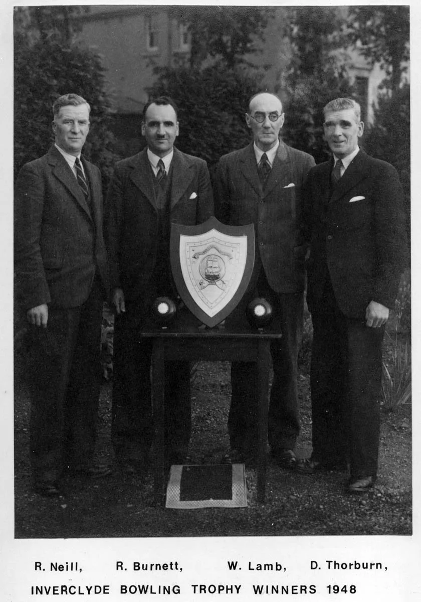 Black and white photo of four men in suits standing outdoors around a table with a shield and two small lights, posing for a photo. The caption reads: R. Neill, R. Burnett, W. Lamb, D. Thorburn, Inverclyde Bowling Trophy Winners 1948.
