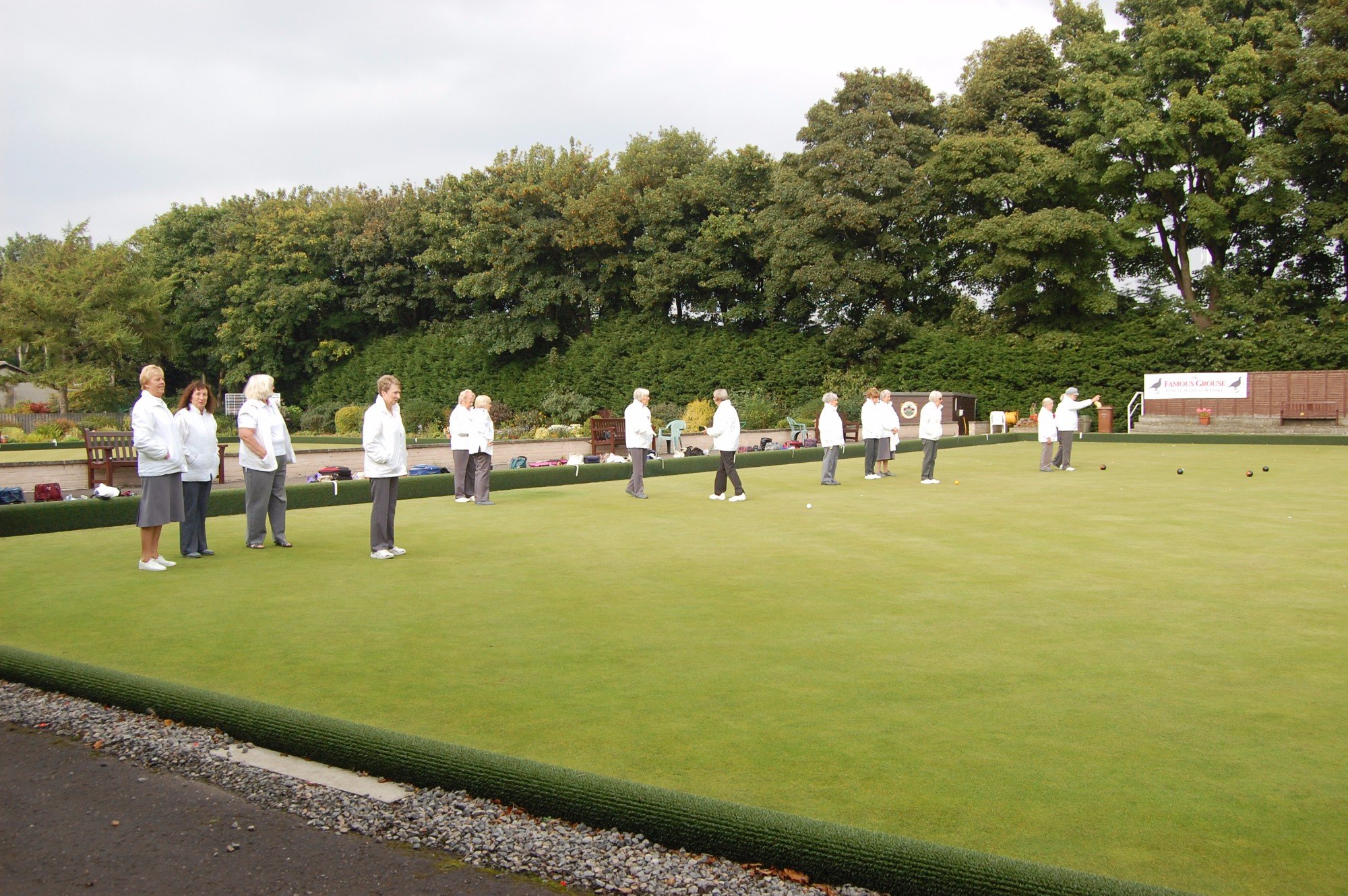 A group of senior women participating in a game of lawn bowls on a well-maintained green, with trees and signage in the background.