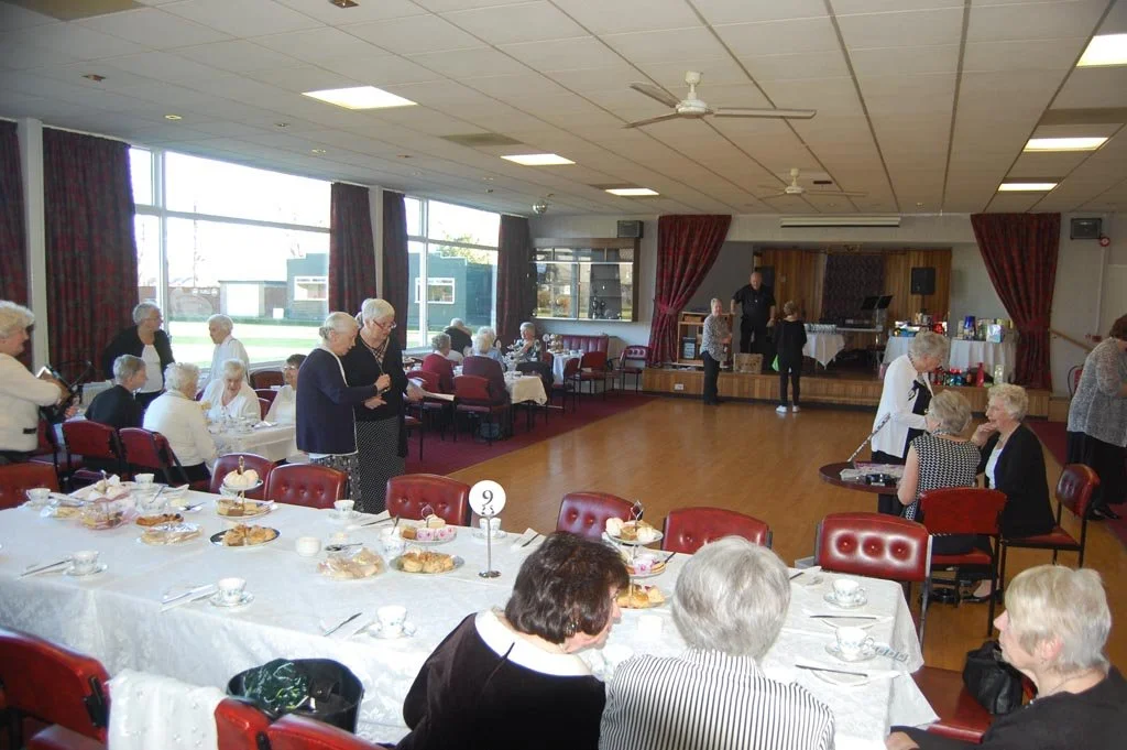 Elderly people gathered in a banquet hall for a social event with a stage, tables set for tea and cake, and a few people standing and sitting.