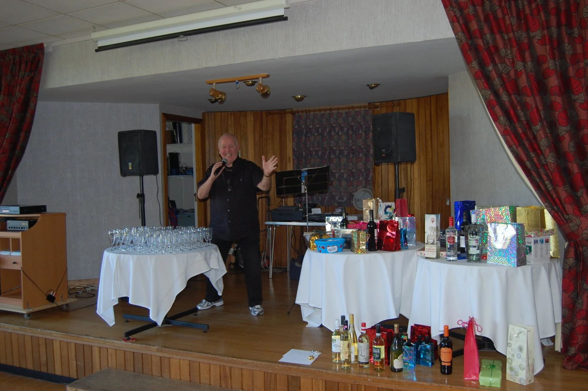 A man holding a microphone, smiling, standing near two tables with wrapped gifts and bottles, in a decorated room with curtains and audio equipment.