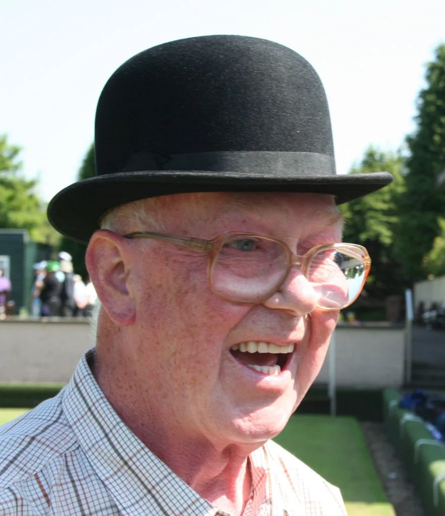 An elderly man wearing a black bowler hat, glasses with a plastic frame, and a checked shirt, smiling outdoors with trees and people in the background.
